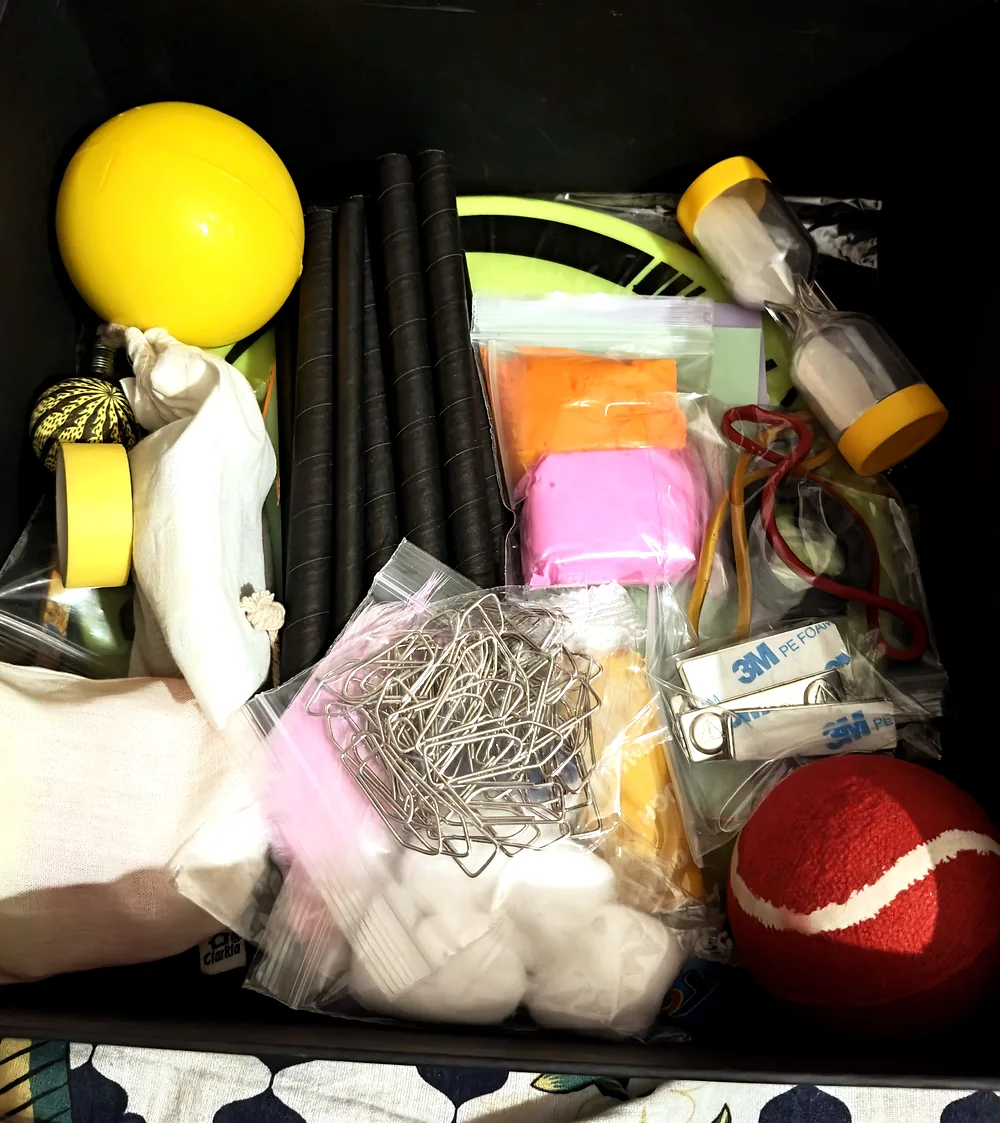 The open EscapeGravity box packed with its real play objects — balls, clay, rods, a frisbee, cotton, rubber bands and more